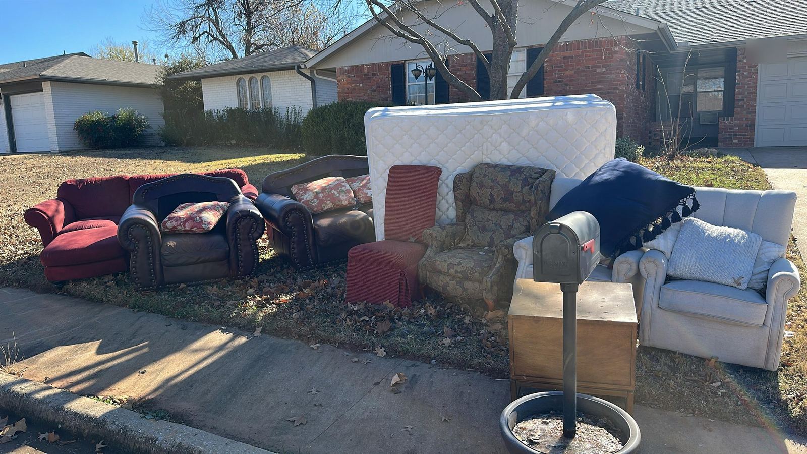 Sofas, armchairs, a mattress and a dresser lined up at the curb in front of a brick home, ready for pickup